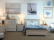 Custom-made bedding furniture display featuring a bed with blue and white linens, matching bedside tables, and decorative lamps, set against a coastal-themed wall art in a showroom environment.